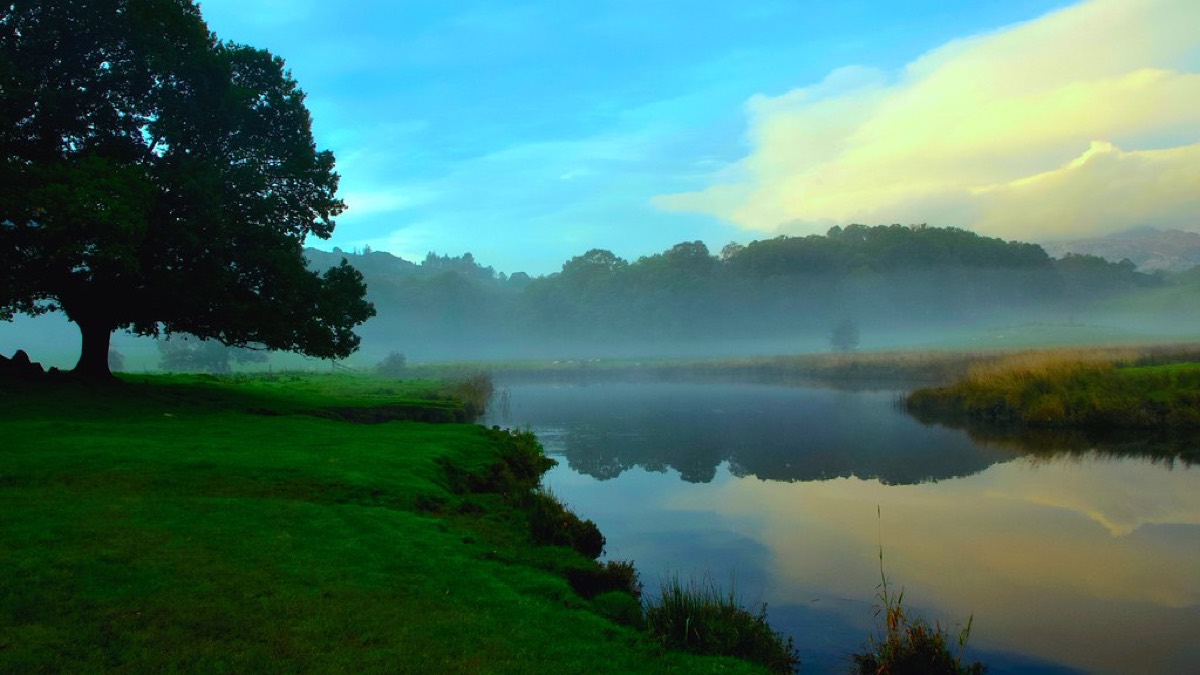 A still river at dawn, mist rising over green banks and distant hills under a soft teal sky.